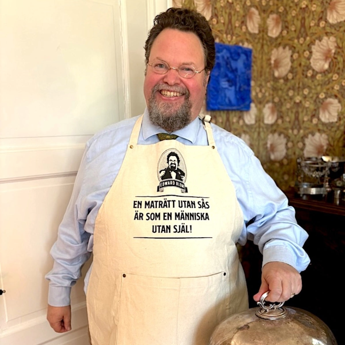 Edward Blom i sitt hem med beige förkläde med citatet "En maträtt utan sås är som en människa utan själ"